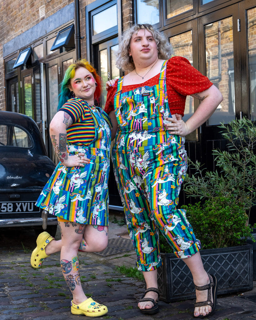 Two femme models pose outside a building wearing vibrant, colourful Run & Fly unicorn-themed outfits. The model on the left has rainbow hair and is in a pinafore dress, while the model on the right has short blonde curly hair and wears dungarees. The unicorn print features rainbows, stars, and cute, cartoon-style unicorns against a striped background. Run & Fly is a small ethical slow fashion company offering quirky, playful designs for all body types.