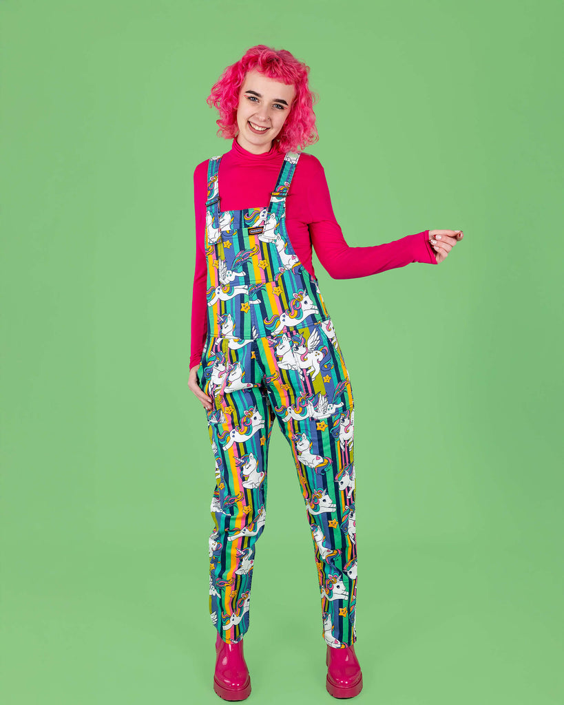 A pink haired tall femme model poses in front of a green background wearing Run & Fly dungarees with a unicorn print. The dungarees feature rainbow-striped backgrounds and white unicorns with wings and rainbow manes. The model pairs the outfit with a bright pink top and pink boots. Run & Fly creates ethical slow fashion, known for bold, colourful prints and inclusive designs for all sizes.