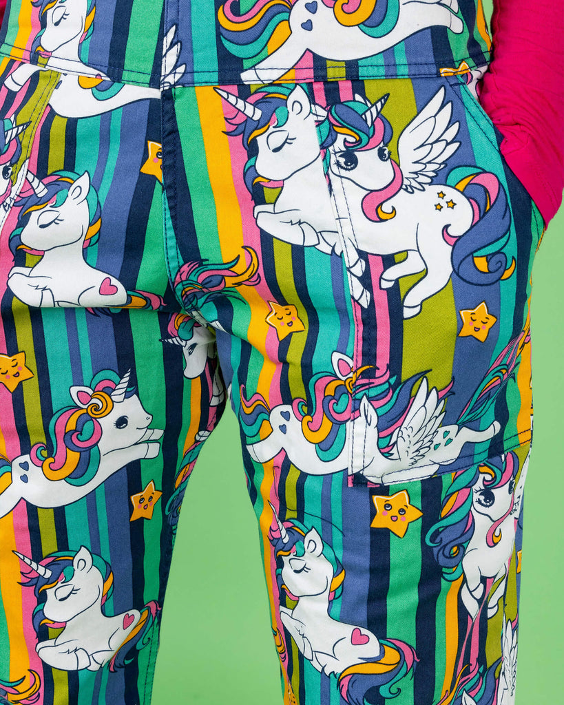 Close-up detail of the Run & Fly dungarees featuring a colourful cartoon unicorn print. The design includes white unicorns with rainbow manes and tails, yellow stars, and vertical stripes in shades of green, blue, pink, and yellow. These playful, eye-catching dungarees are part of Run & Fly’s ethical, slow fashion range, showcasing vibrant and fun styles.