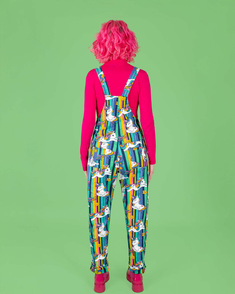 Rear view of a pink haired tall femme model showing the back of the Run & Fly unicorn print dungarees. The dungarees have rainbow-striped backgrounds with white unicorns and stars in a cartoon-style print. The bright, whimsical design is paired with a bold pink long-sleeve top. Run & Fly is a small ethical slow fashion brand, known for creating unique and colourful clothing for all shapes and sizes.