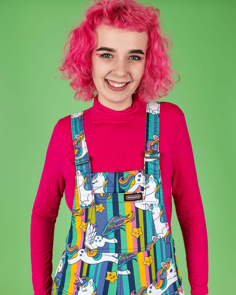 A pink haired tall femme smiling model poses in front of a green backdrop, showing off the vibrant Run & Fly unicorn dungarees. The dungarees feature a playful pattern with rainbow-striped backgrounds and cute cartoon unicorns. The model is wearing a bright pink top, which enhances the bold colours of the design. Run & Fly focuses on quirky, ethical slow fashion with standout prints.