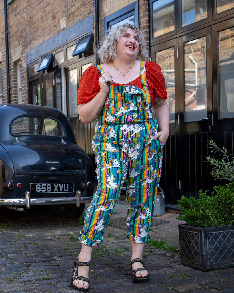 A laughing femme blonde hair model wearing Run & Fly’s unicorn print dungarees, which feature rainbow-striped backgrounds and cute unicorns with stars. The dungarees are styled with a red puff sleeve top and sandals. The model stands outside on a cobblestone street, radiating joy. Run & Fly specialises in bold, inclusive slow fashion for all body types with standout prints.