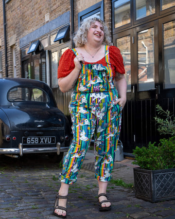A laughing femme blonde hair model wearing Run & Fly’s unicorn print dungarees, which feature rainbow-striped backgrounds and cute unicorns with stars. The dungarees are styled with a red puff sleeve top and sandals. The model stands outside on a cobblestone street, radiating joy. Run & Fly specialises in bold, inclusive slow fashion for all body types with standout prints.