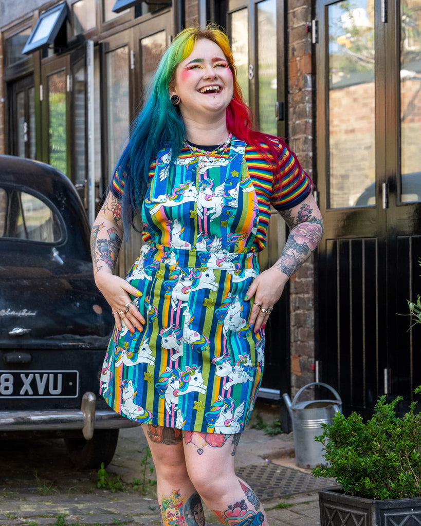 A model with rainbow-coloured hair and colourful makeup stood outside, wearing Unicorn Stripes Stretch Twill Pinafore Dress from ethical, slow fashion brand Run & Fly, paired with a rainbow striped t shirt.They are smiling and posing with their arms out by their sides, and visible tattoos on their arms and legs.