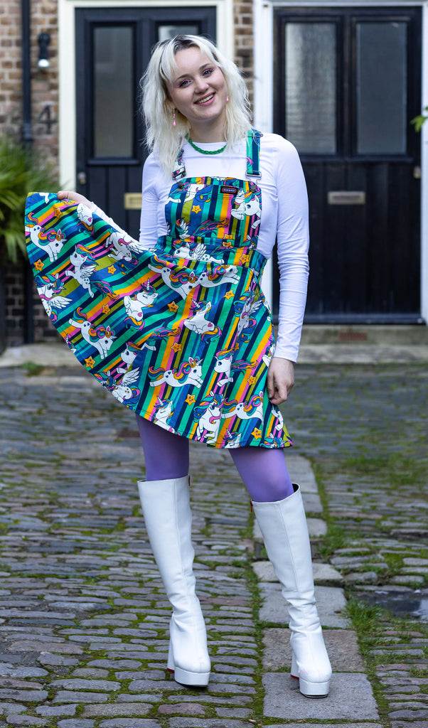 A blonde femme model wearing the unicorn stripes flared pinafore dress from run & fly with a long sleeve white top, purple tights and white platform boots. They are outside in a courtyard facing forward smiling whilst holding out the bottom of the pinafore. The slow fashion pinafore dress features rainbow stripes, kawaii cute pegasus', unicorns and stars with faces.