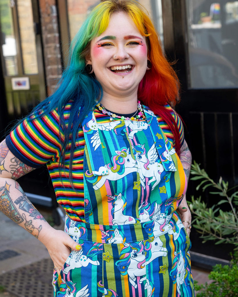 Close up of a model with rainbow-coloured hair and vibrant makeup wearing Run & Fly Unicorn Stripes Stretch Twill Pinafore Dress paired with a rainbow striped t shirt underneath. They are smiling with their hands in the pockets of the dress.  