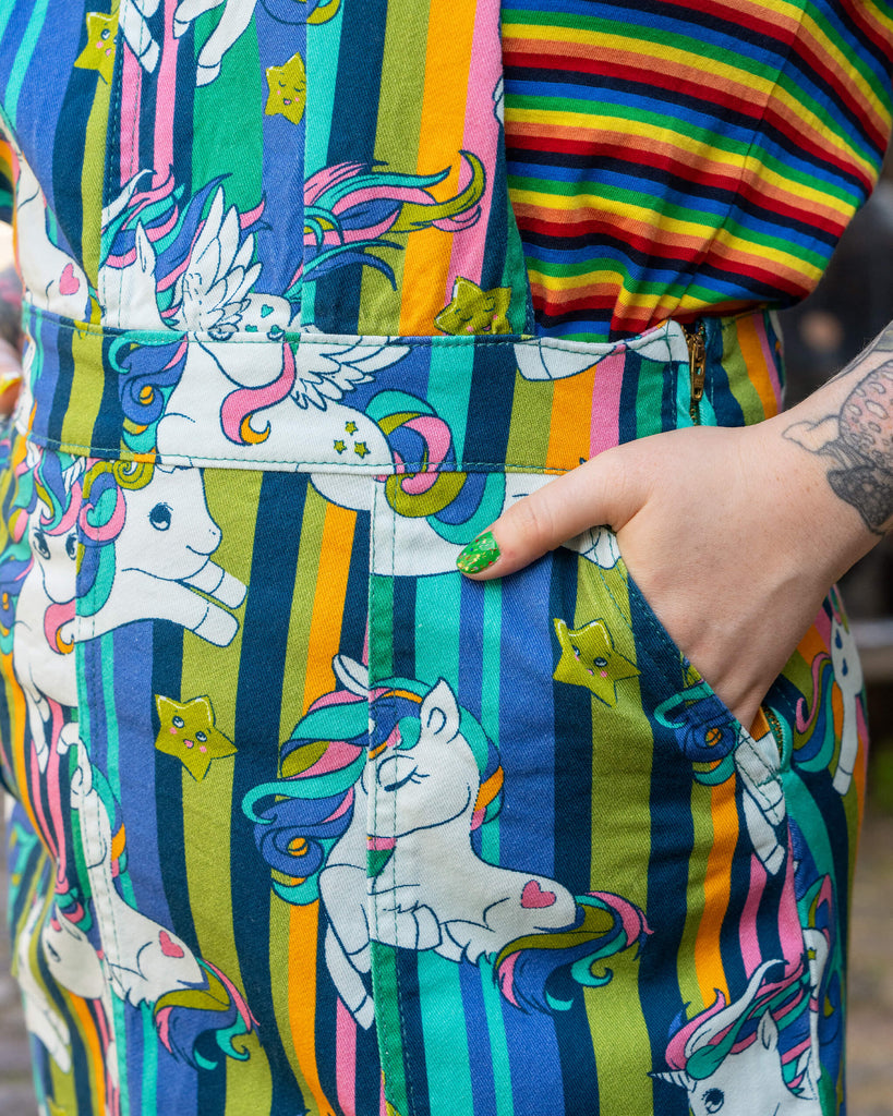 Close up of a model with rainbow-coloured hair and vibrant makeup wearing Run & Fly Unicorn Stripes Stretch Twill Pinafore Dress paired with a rainbow striped t shirt underneath. They are smiling with their hands in the pockets of the dress.  