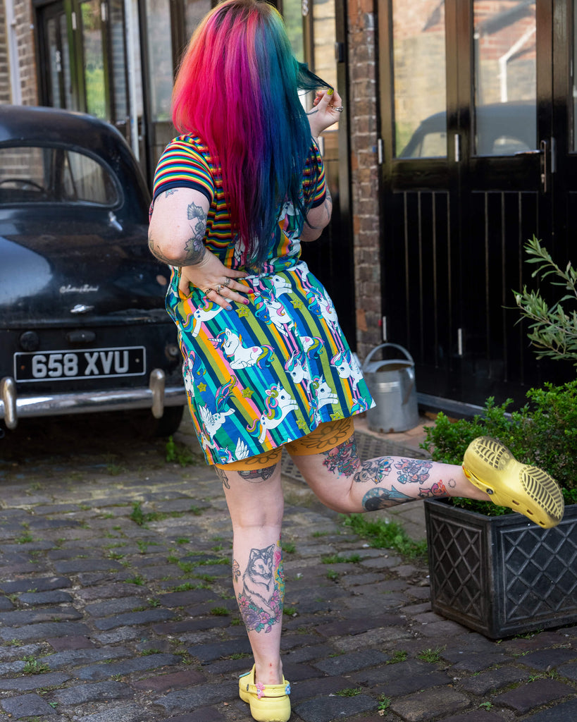 A person with rainbow-coloured hair facing away from the camera, showcasing the back of their unicorn print stretch twill pinafore dress from Run & Fly. They are standing on a cobbled street, with visible tattoos on their arms and legs, and yellow Crocs.