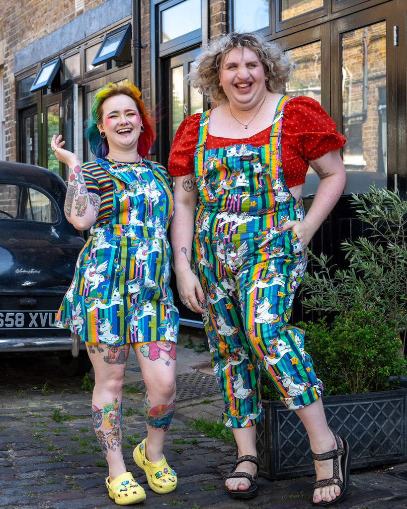 Two models smile brightly while wearing Run & Fly unicorn-themed outfits. The model on the left sports a colourful pinafore dress with cartoon unicorns and rainbow-striped backgrounds, paired with yellow Crocs. The model on the right wears dungarees with the same playful unicorn print, paired with a red puff sleeve top and sandals. Run & Fly is a small ethical slow fashion company known for inclusive, bold, and quirky designs that fit all body types.