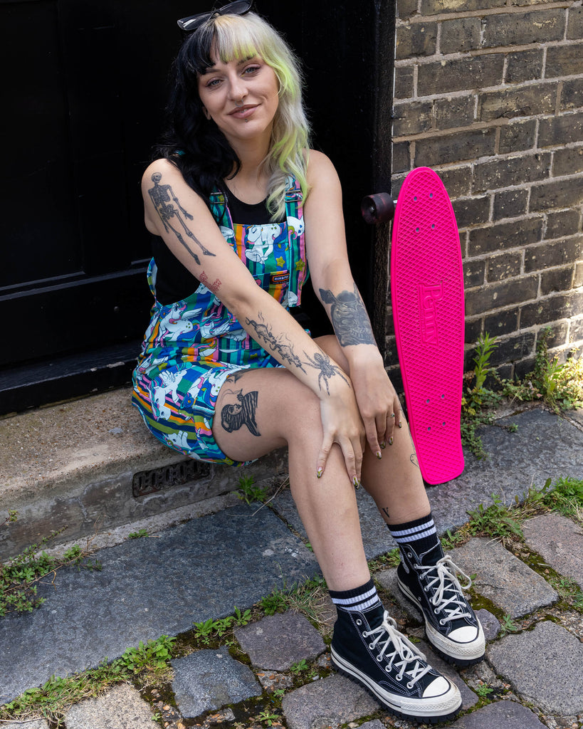 The woman sits on a step outside, smiling with her oversized sunglasses on her head. She is wearing a black tank top under her vibrant unicorn print short dungarees by Run & Fly, and black high-top Converse trainers. Her bright pink Penny skateboard is nearby.