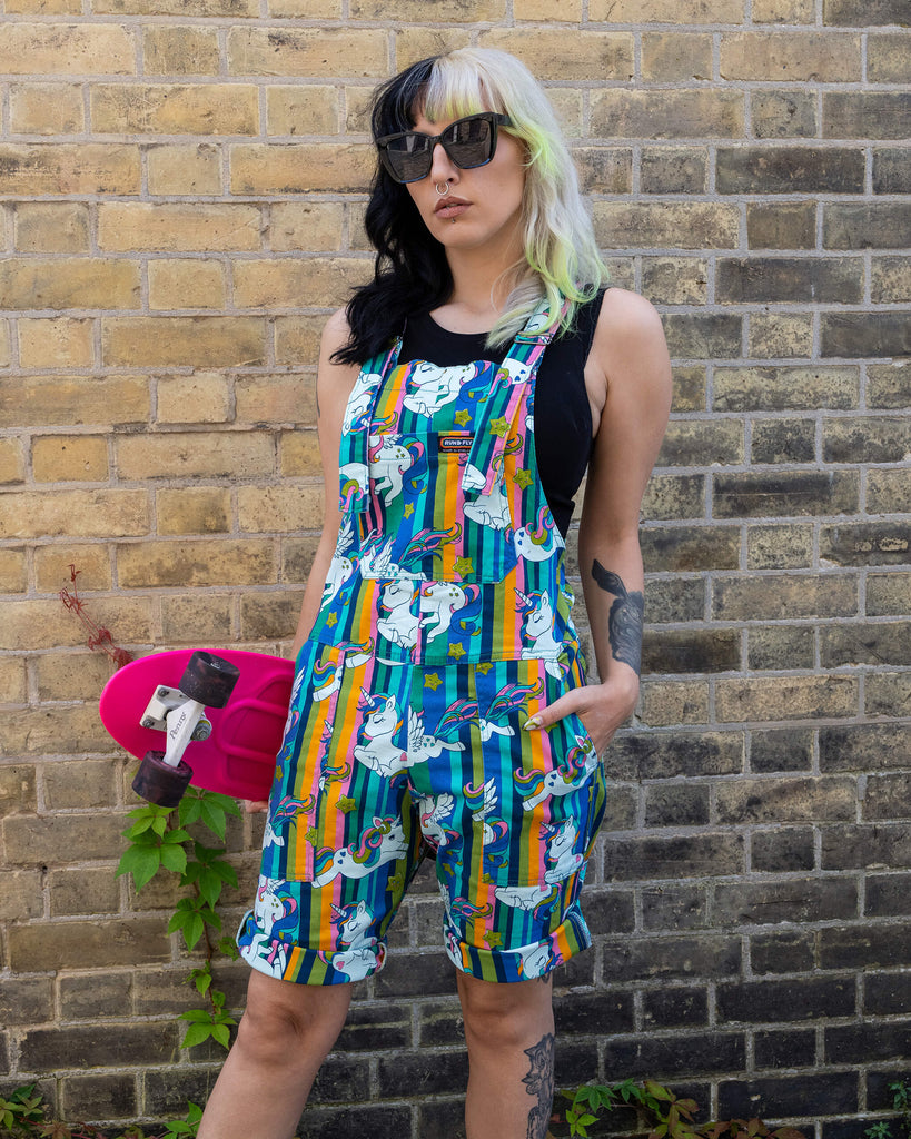 The woman stands confidently against a brick wall, one hand in the pocket of her unicorn print short dungarees by Run & Fly. She wears black high-top Converse trainers and a black tank top. Her bright pink Penny skateboard is held in one hand.