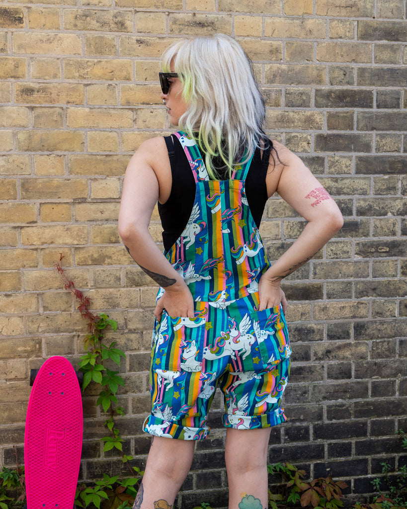 Standing against a brick wall, the woman models the back view of her colourful unicorn print short dungarees by Run & Fly. Her hands are on her hips, and her bright pink Penny skateboard is leaning against the wall beside her.