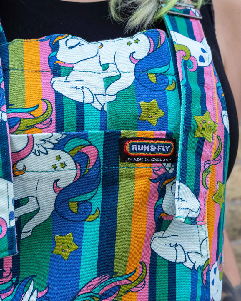 Close-up of the unicorn print short dungarees by Run & Fly, showing the detailed and colourful pattern. The background is blurred, focusing on the vibrant design of the dungarees.