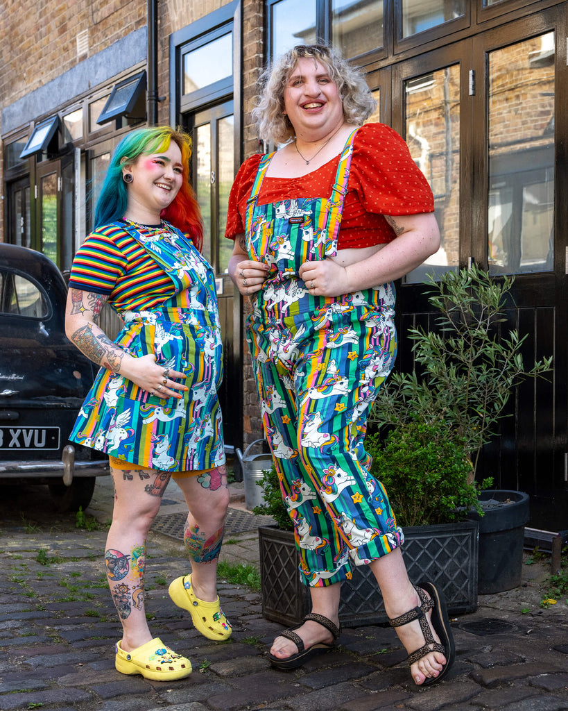 Two friends laughing and striking poses outdoors, the model on the left is wearing unicorn stripes stretch twill pinafore dress paired with rainbow striped t shirt from Run & Fly, the other wearing unicorn stripes stretch twill dungarees from Run & Fly paired with a red polka dot top.