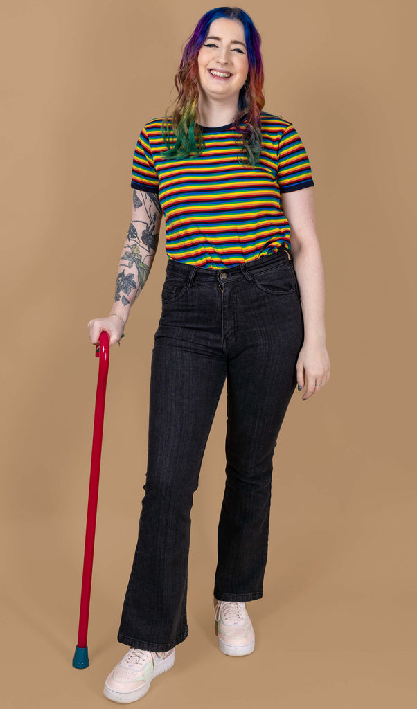 Eliza is stood wearing the Unisex Black Stonewashed Bellbottom Flares Leg High Waisted Jeans with the rainbow stripe short sleeve tshirt and pastel pink trainers. They have rainbow hair, nature themed tattoos on one arm and a red walking aid whilst smiling to camera. The jeans are high waisted with a faded stonewash black colour, fitted to thigh and flared bottoms.