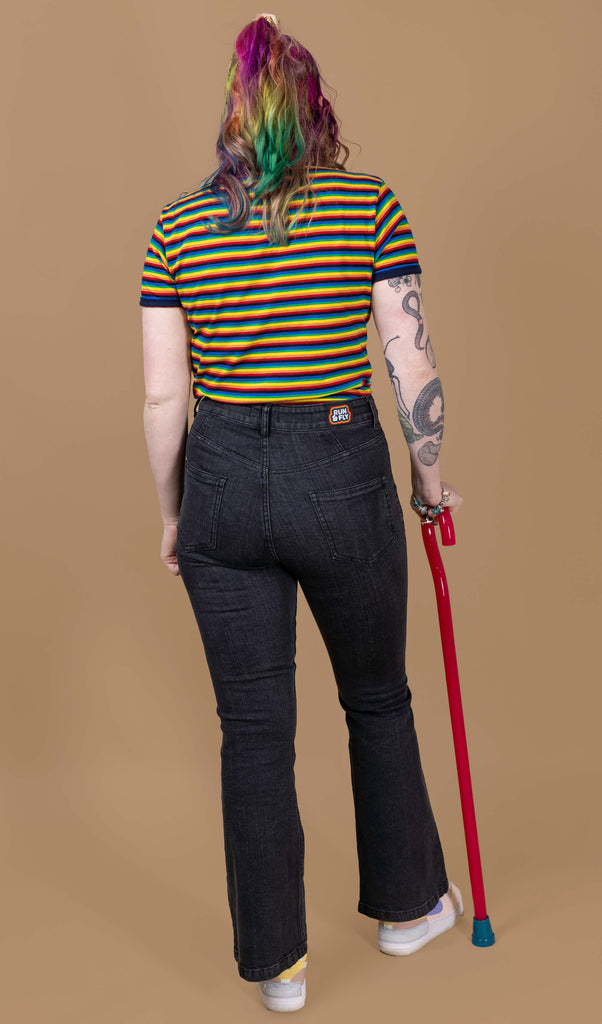 Eliza is stood wearing the Unisex Black Stonewashed Bellbottom Flares Leg High Waisted Jeans with the rainbow stripe short sleeve tshirt and pastel pink trainers. They have rainbow hair, nature themed tattoos on one arm and a red walking aid whilst smiling to camera. The jeans are high waisted with a faded stonewash black colour, fitted to thigh and flared bottoms.