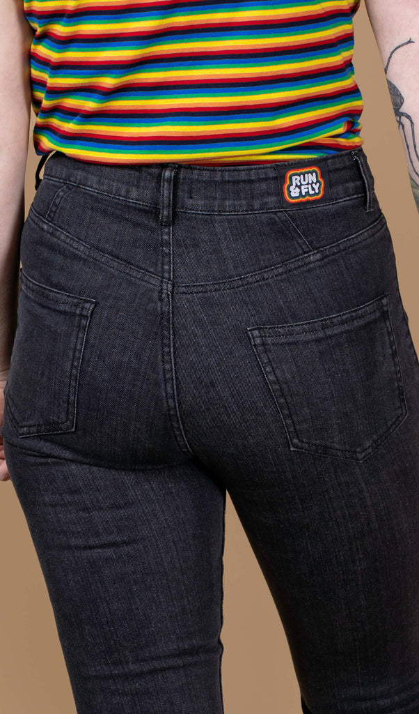 Close up of the back of Eliza stood wearing the Unisex Black Stonewashed Bellbottom Flares Leg High Waisted Jeans with the rainbow stripe short sleeve tshirt. They are stood with one arm resting by their side whilst the other holding the walking aid. The jeans are high waisted with a faded stonewash black colour, fitted to thigh and flared bottoms.