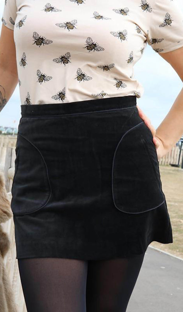 Uk size 10 model wearing a Run and Fly Black Velvet A-Line Mini Skirt in size M. The photo has been cropped to the torso and hips. The model has one hand on their hip and wearing a matching cream bumblebee print, short-sleeved t-shirt.