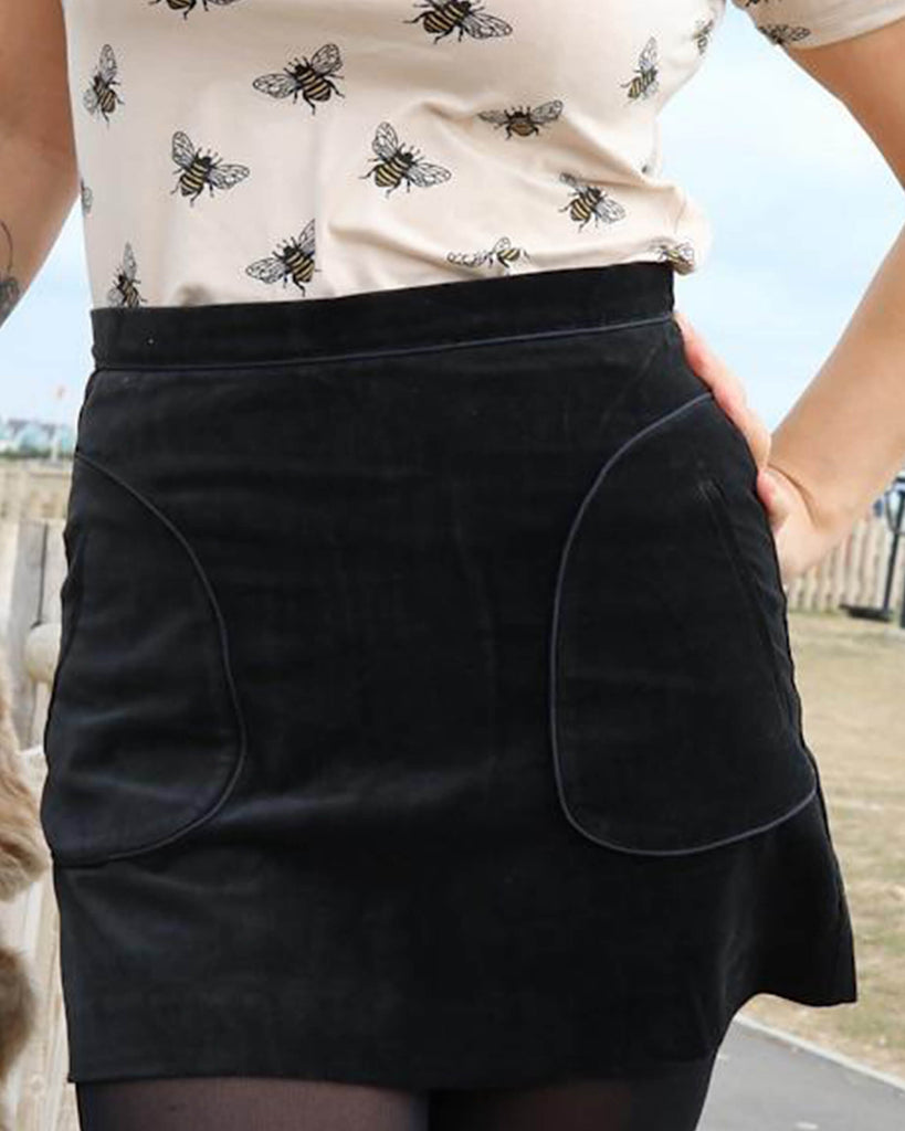 Uk size 10 model wearing a Run and Fly Black Velvet A-Line Mini Skirt in size M. The photo has been cropped to the torso and hips. The model has one hand on their hip and wearing a matching cream bumblebee print, short-sleeved t-shirt.