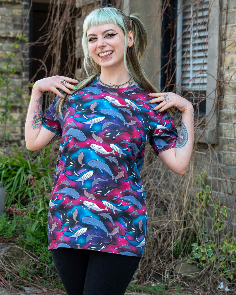 Person posing joyfully in the Run & Fly Cosmic Sea T Shirt, with an all-over print of sea creatures like dolphins and whales on a bright galaxy-style background. A colourful, comfy, short sleeve top from a small UK ethical fashion brand.