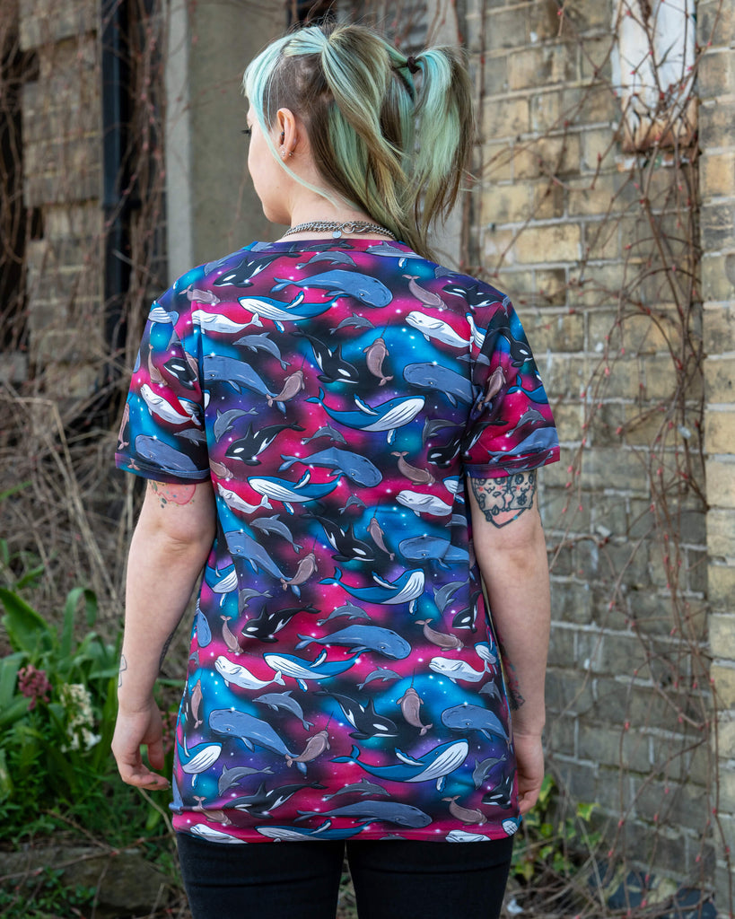 Full back view of a person standing outside wearing the Run & Fly Cosmic Sea T Shirt, featuring a vibrant ocean creature print on a galactic background. The short sleeve top showcases narwhals, whales, and dolphins in a rainbow nebula-style design. Ethically made in England.