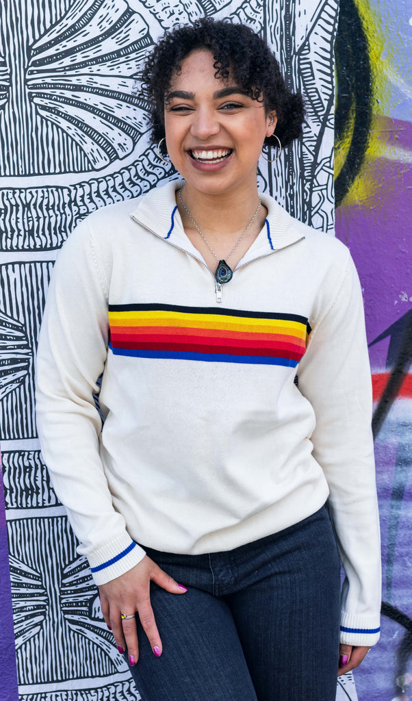 Georgia is stood in front of a graffiti wall wearing the ecru retro zip neck jumper with denim jeans. They are facing forward posing with one hand on their leg and the other hand resting by their side whilst laughing to camera. Photo is cropped from the thighs up.