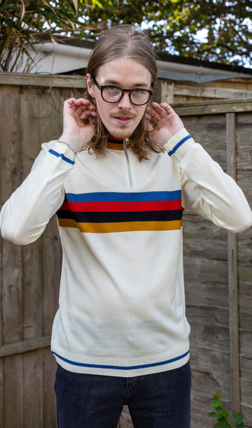 Jack a white male with shoulder length blonde hair and glasses is wearing a high neck knit jumpers with stripes across the middle. He has his hands in his hair and is in a garden