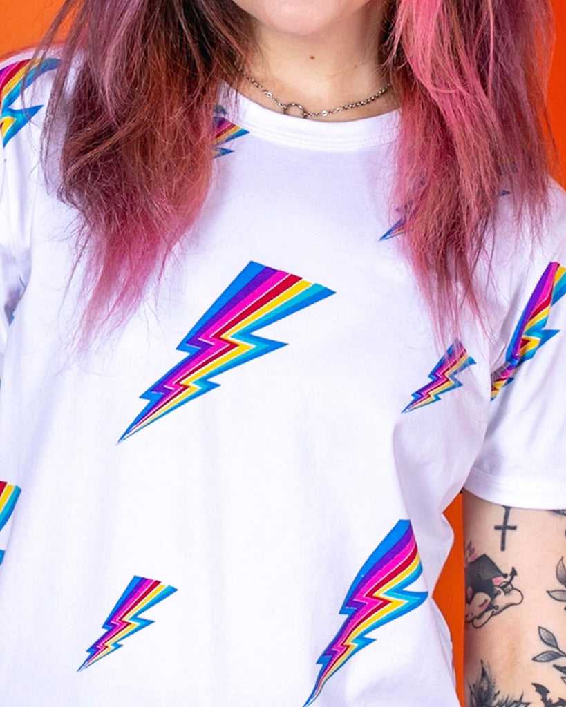 Close up of Sarah wearing the white lightning bolt short sleeve tee. She has pink and brown hair in bunches with pink makeup and arm tattoos. The tshirt is a white base colour with repeating rainbow lightning bolts varying in size all over.