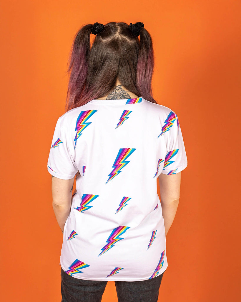 Sarah is stood wearing the white lightning bolt short sleeve tee with black stonewash denim flares. She has pink and brown hair in bunches with pink makeup and arm tattoos. She is facing away from the camera with both arms resting in front of her. The tshirt is a white base colour with repeating rainbow lightning bolts varying in size all over.