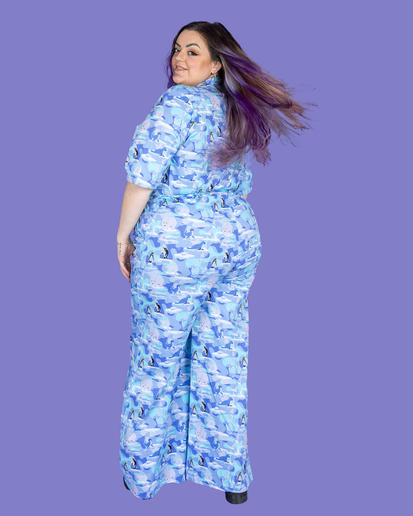 a model with purple hair wearing Winter Friends Stretch Jumpsuit paired with black boots. The jumpsuit is a light blue colour with ice burgs, seals, polar bears and penguins on. Model is standing with their back to the camera and looking back over their shoulder with their hair swishing. The background of the photo is purple.