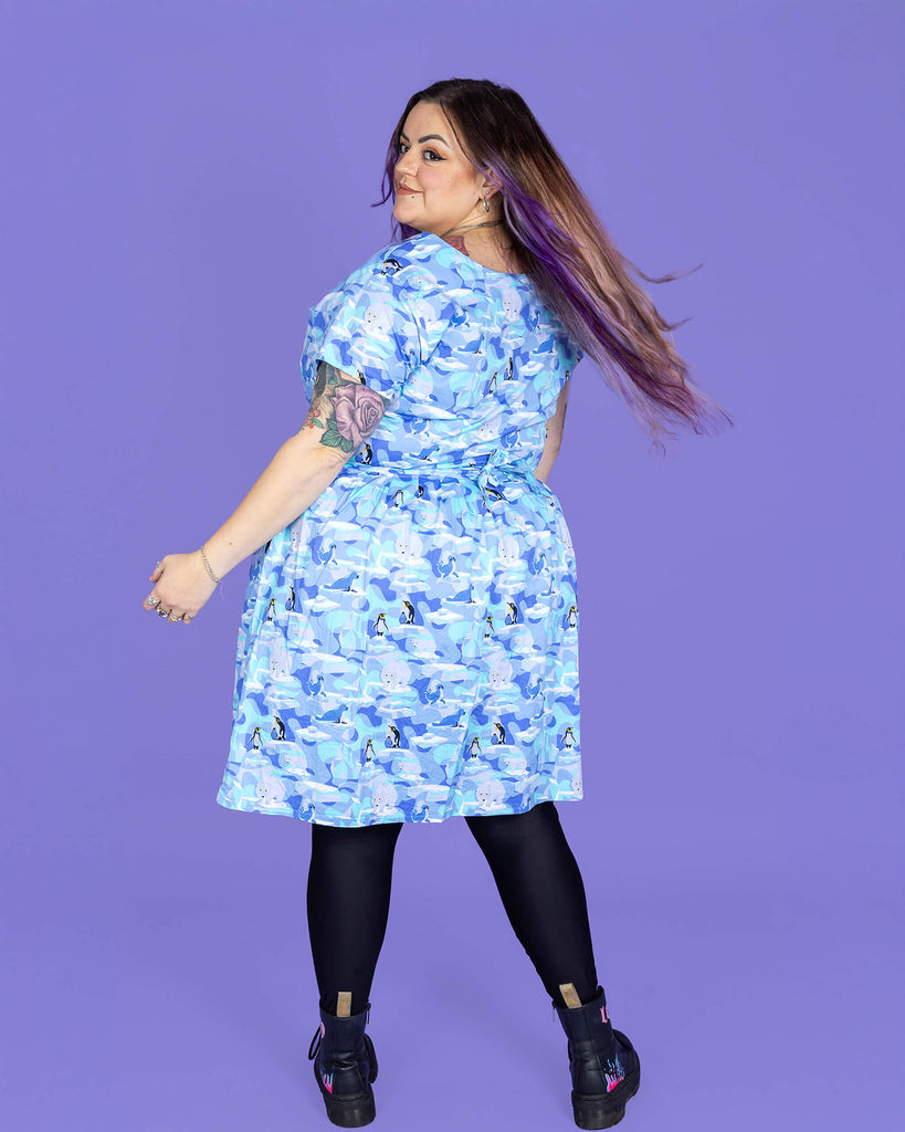 a model with purple hair wearing Winter Friends Stretch Belted Tea Dress with Pockets paired with black boots and tights. The jumpsuit is a light blue colour with ice burgs, seals, polar bears and penguins on. The model is standing with their back to the camera while looking back over their shoulder and swishing their hair in front of a purple background.
