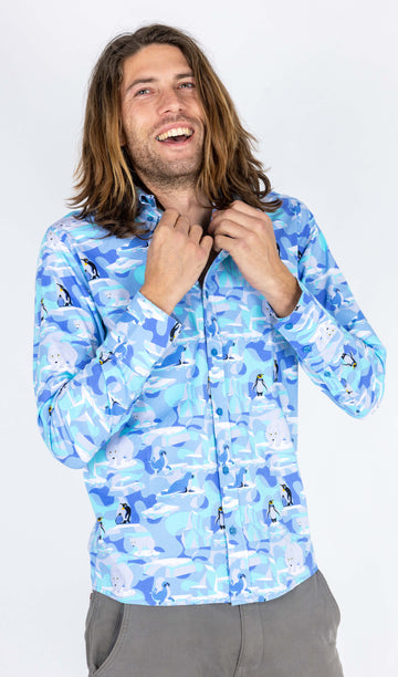 Rudi a male model with shoulder length hair is wearing Run & Fly Winter Friends Long Sleeve Shirt paired with grey trousers. The shirt is ight blue colour with ice burgs, seals, polar bears and penguins on. Rudi is smiling at the camera and holding the collar of the shirt in front of a white background.
