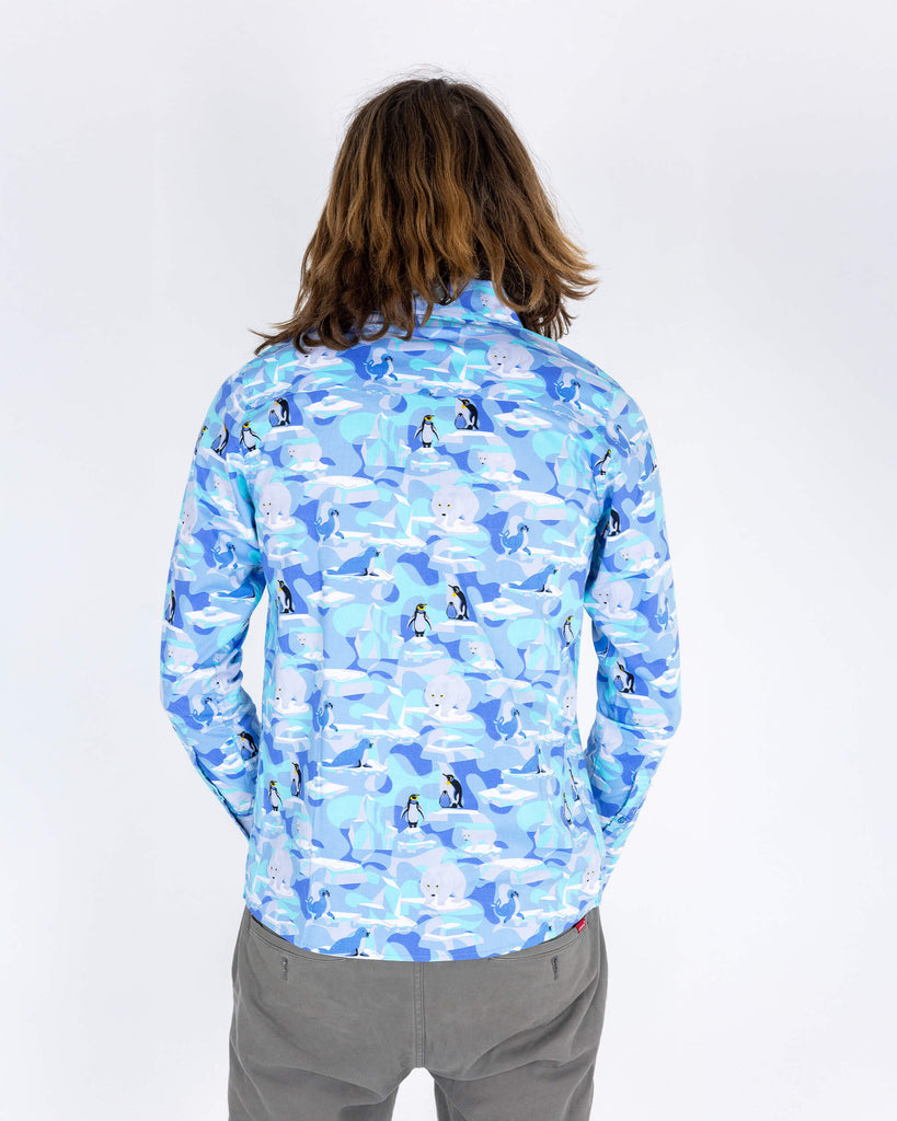 Rudi a male model with shoulder length hair is wearing Run & Fly Winter Friends Long Sleeve Shirt paired with grey trousers. The shirt is light blue colour with ice burgs, seals, polar bears and penguins on. Rudi is stood with his back to the camera in front of a white background.