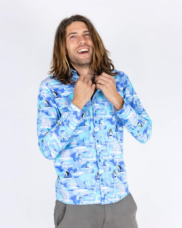 Rudi a male model with shoulder length hair is wearing Run & Fly Winter Friends Long Sleeve Shirt paired with grey trousers. The shirt is light blue colour with ice burgs, seals, polar bears and penguins on. Rudi is posing with hands on the collar 