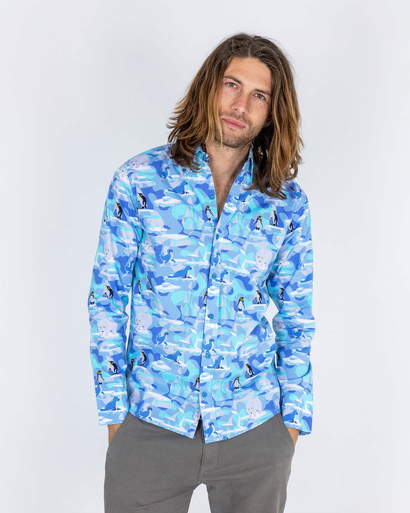 Rudi a male model with shoulder length hair is wearing Run & Fly Winter Friends Long Sleeve Shirt paired with grey trousers. The shirt is light blue colour with ice burgs, seals, polar bears and penguins on. Rudi is posing with hands in his pockets in front of a white background.