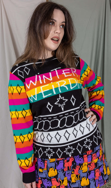 Alice is stood in front of a draped sheet background wearing the Run & Fly x Katie Abey Winter Weirdo Jumper with cats club pinafore underneath. She is facing forward with one hand on her hip, the other hand by her side whilst looking off to the right. The jumper has rainbow sleeves with black cuffs and sparkles, the chest is black and white christmas sparkles and patterns striped down it with 'Winter Weirdo' written in the centre with a rainbow behind the text to match the sleeves.