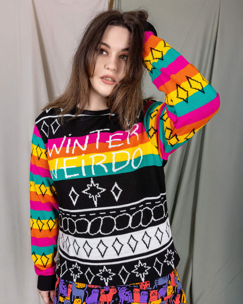 Alice is stood in front of a draped sheet background wearing the Run & Fly x Katie Abey Winter Weirdo Jumper with cats club pinafore underneath. She is facing forward with one hand on her head, the other hand by her side whilst looking to the left. The jumper has rainbow sleeves with black cuffs and sparkles, the chest is black and white christmas sparkles and patterns striped down it with 'Winter Weirdo' written in the centre with a rainbow behind the text to match the sleeves.
