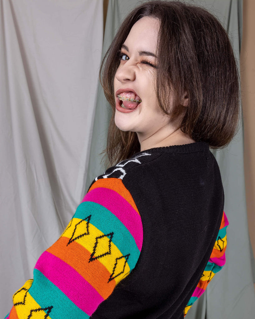 Alice is stood in front of a draped sheet background wearing the Run & Fly x Katie Abey Winter Weirdo Jumper with cats club pinafore underneath. She is facing away looking over her shoulder sticking her tongue out. The jumper has rainbow sleeves with black cuffs and sparkles, the back middle is plain black. Photo is cropped from the hips up.