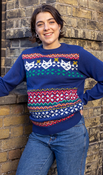 Freya is a white woman in her 20's with short brown hair wearing a blue christmas style jumper with cats on. She is leaning on a brick wall