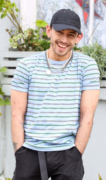 Zach is stood in front of a house wearing the retro sky marl pinstriped tshirt with black trousers. He's paired it with a pearl necklace, a safety pin chain and black cap. He's facing forward posing with both hands in his front trouser pockets whilst smiling. Photo is cropped from the hips up.