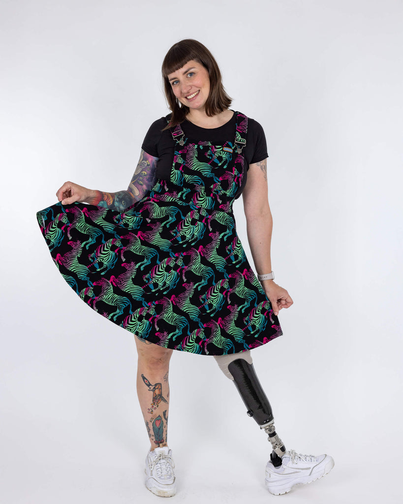 Laura, a white femme model with tattoos, is wearing Rainbow Zebra Flared Pinafore Dress with a black t-shirt underneath. The black pinafore dress has an all over colourful zebra print. Laura is stood in front of a white backdrop at a photography studio in Hove smiling facing the camera with one leg turned out and holding the skirt of the dress. 