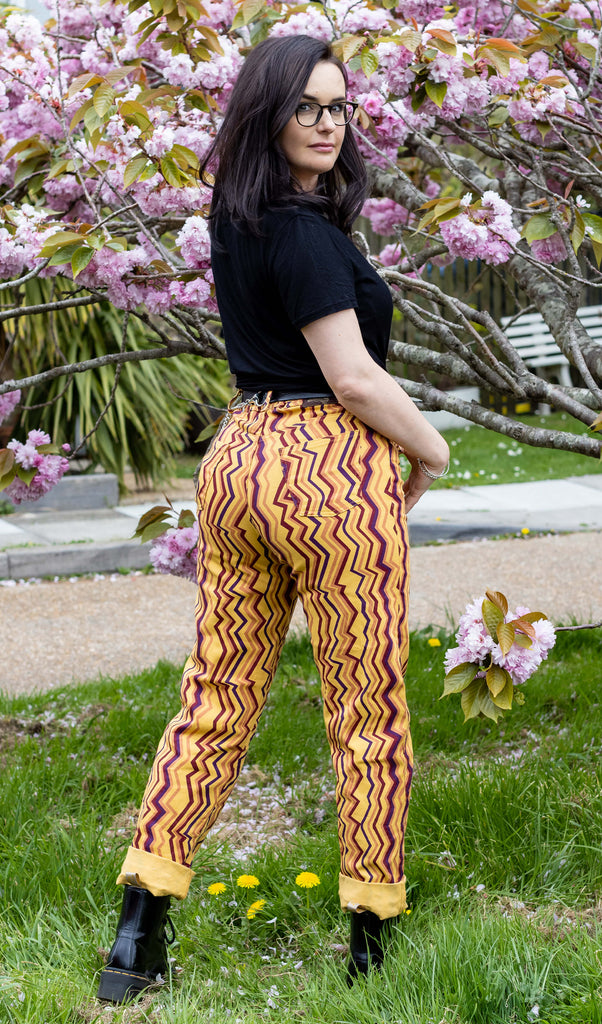 Amy is wearing the 90's zigzag straight leg high waisted jeans with a long sleeve black tshirt and yellow black boots looking over their shoulder in front of a blossom tree