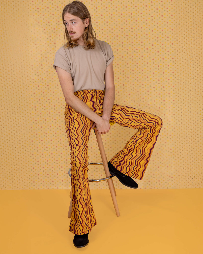 Jack is sat on a stool in front of a yellow studio background wearing the zig zag print bell bottom stretch super flares with a beige short sleeved tshirt and black boots. They are sat with one leg up on the stool foot rest to highlight the flares and both hands resting in the middle of their lap whilst looking to camera.