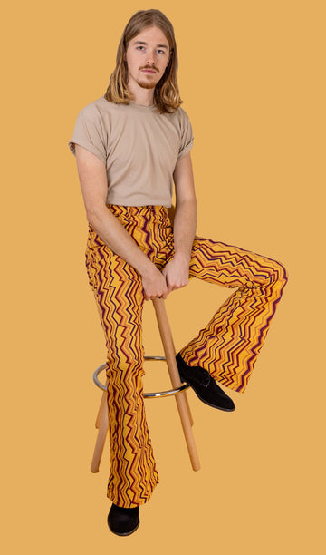 Jack is sat on a stool in front of a yellow studio background wearing the zig zag print bell bottom stretch super flares with a beige short sleeved tshirt and black boots. They are sat with one leg up on the stool foot rest to highlight the flares and both hands resting in the middle of their lap whilst looking to camera.