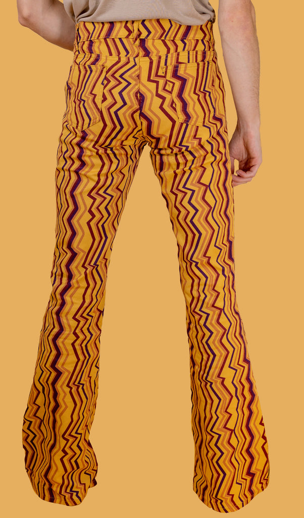 Jack is stood in front of a yellow studio background wearing the zig zag print bell bottom stretch super flares with a beige short sleeve tshirt and black boots. They are facing away from the camera with one hand resting by their side. Photo is cropped from the waist down.