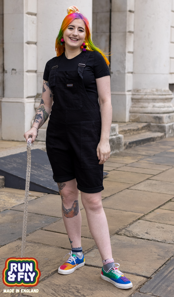 Eliza with a walking stick, laughing and wearing the Black Denim Stretch Dungaree Shorts with a black tee and multicolour trainers