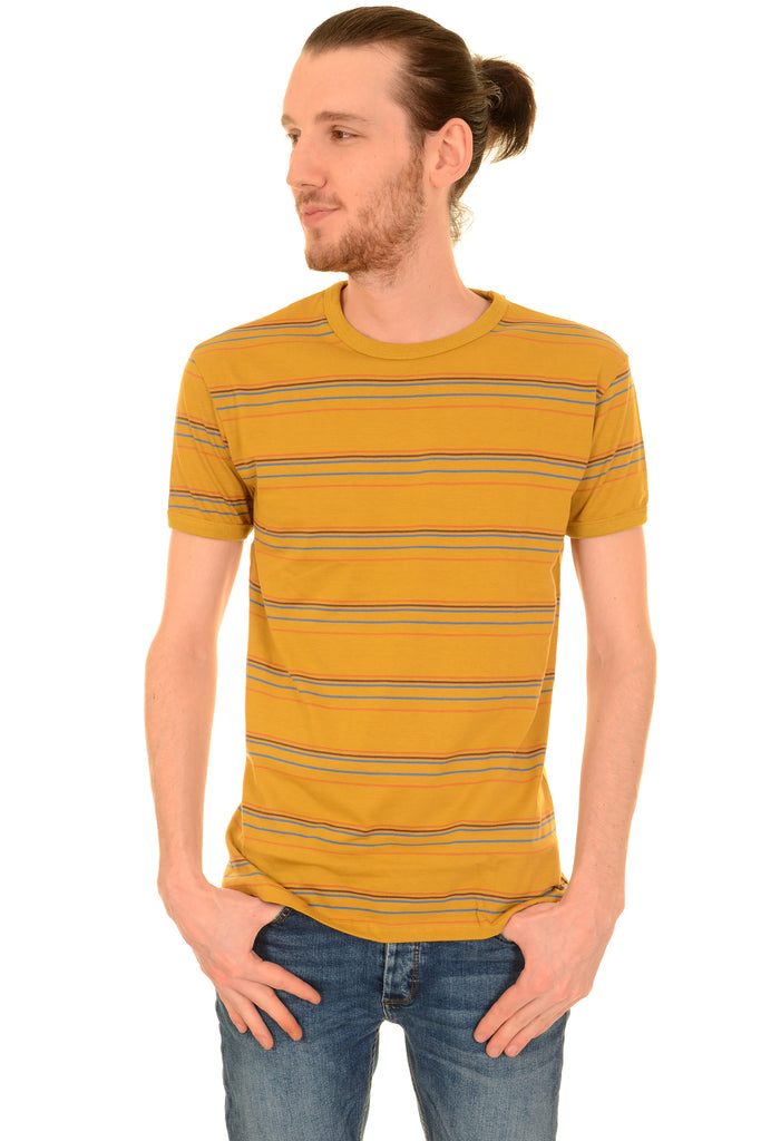 Model is standing in front of a white studio background wearing the sunset stripe retro tshirt with blue jeans. They are facing forward posing with both hands tucked in their jean pockets and looking to the left. Photo is cropped from the knees up.