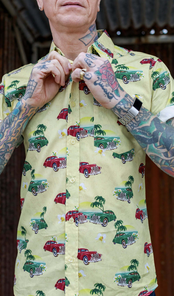 A heavily tattooed model is stood in front of a rusted metal wall wearing the Cuba short sleeve shirt with denim jeans. They are facing the camera with both hands adjusting the top button. Photo is cropped from the lips to hips.