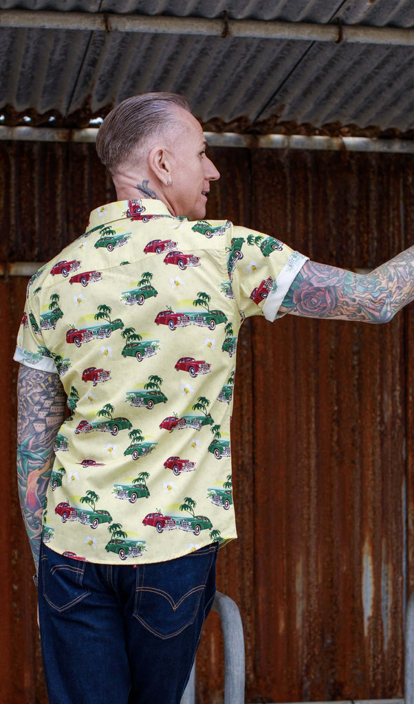 A heavily tattooed model is stood in front of a rusted metal wall wearing the Cuba short sleeve shirt with denim jeans. They are facing away from the camera with one hand resting up on the wall and the other by their side. They are looking over their shoulder to the right. Photo is cropped from the knees up.