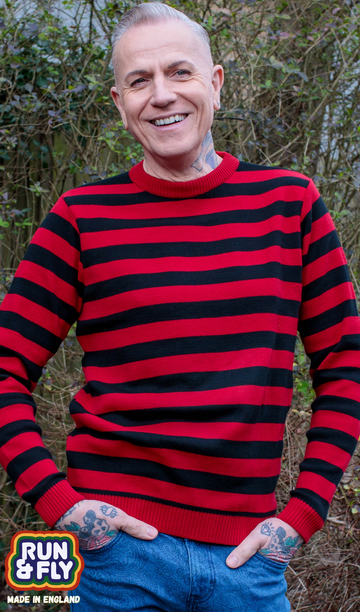 Jim is stood outside in a park area wearing the red and black striped jumper with blue denim jeans. They are heavily tattooed with swept back silver hair. They are facing the camera posing with both hands in their front jean pockets and smiling. Photo is cropped from the hips up.